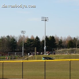 A sports field with a group of people engaging in athletic activities in the background. There is sports equipment like a soccer goal and a scoreboard visible. A chain-link fence runs across the foreground, and a statue or sculpture resembling an eagle is also seen. Trees and tall floodlights are positioned behind the field.