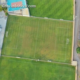 An aerial view of a sports complex featuring multiple green grass fields, including football pitches with marked goalposts. Adjacent to the fields is a blue-roofed building and a smaller area with a tennis or basketball court. Surrounding the complex, there's a road lined with trees and a dirt area with sparse vegetation.